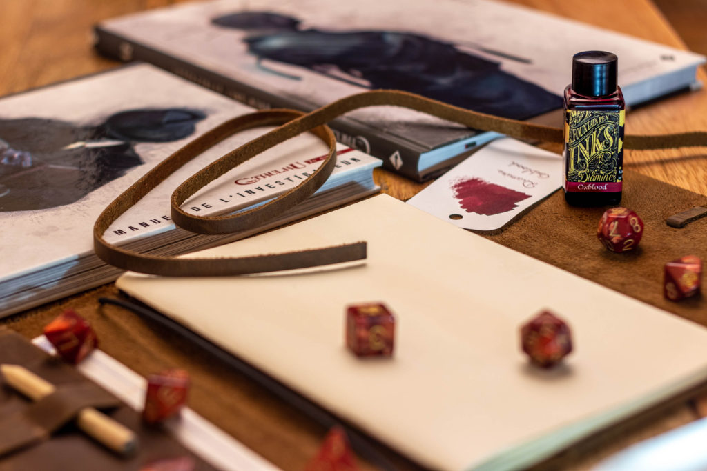 L'encre Diamine Oxblood, sur un carnet de note en cuir, avec des dés et des livres de jeu de rôle.